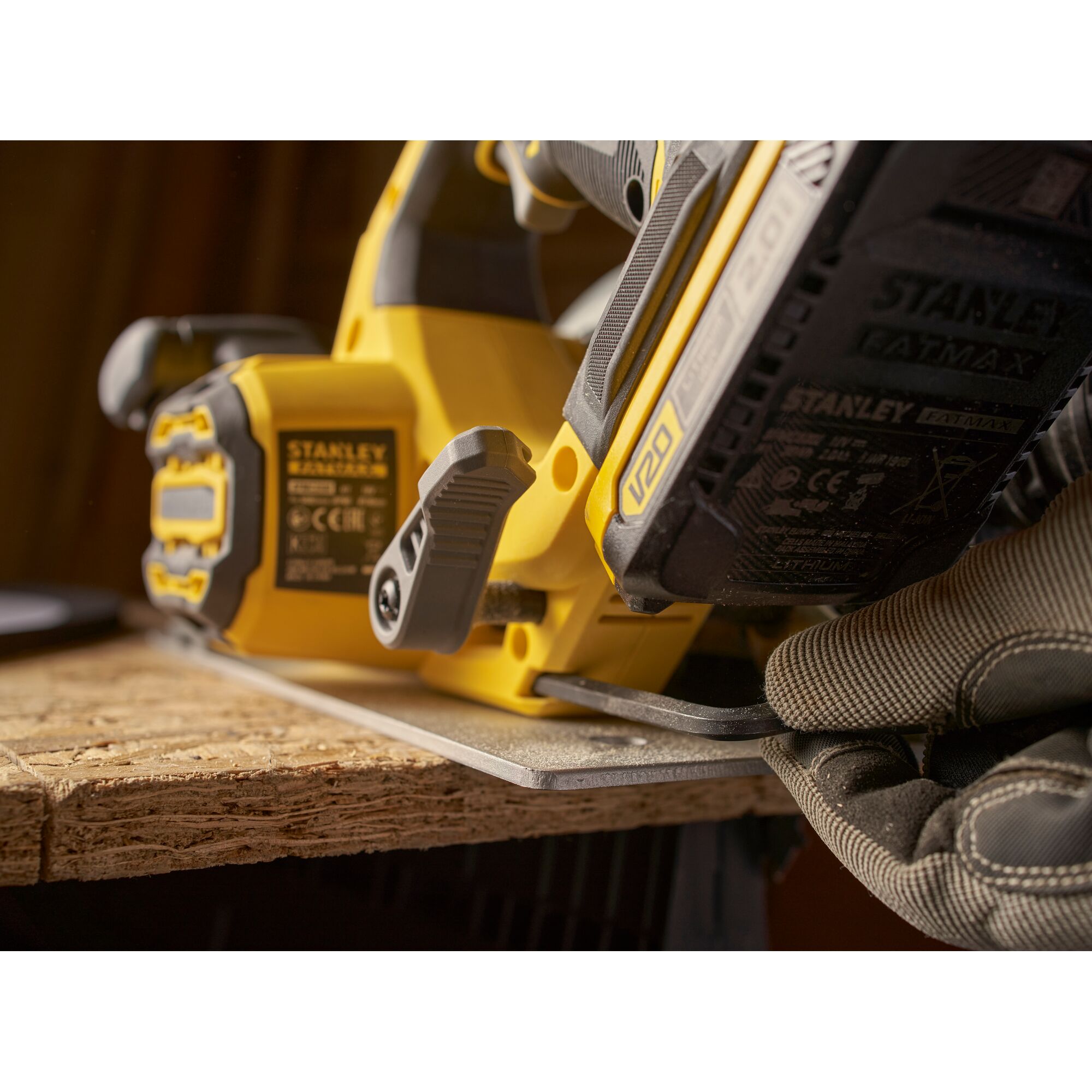 Stanley SBC550 power tool close-up, yellow cordless saw cutting wood, gloved hand adjusts base.