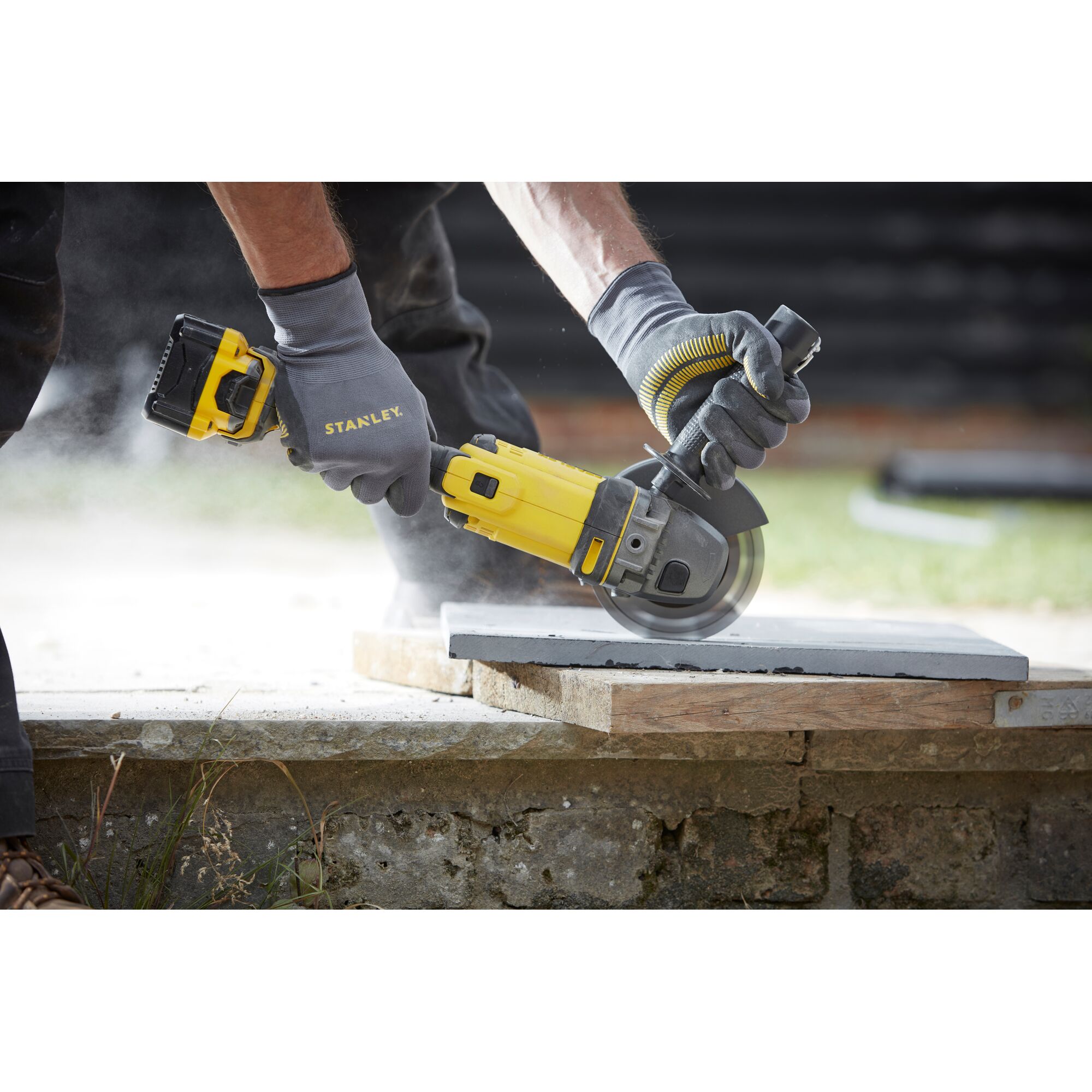 Photo of STANLEY SCG400 cordless angle grinder cutting a concrete slab outdoors, held by gloved hands.
