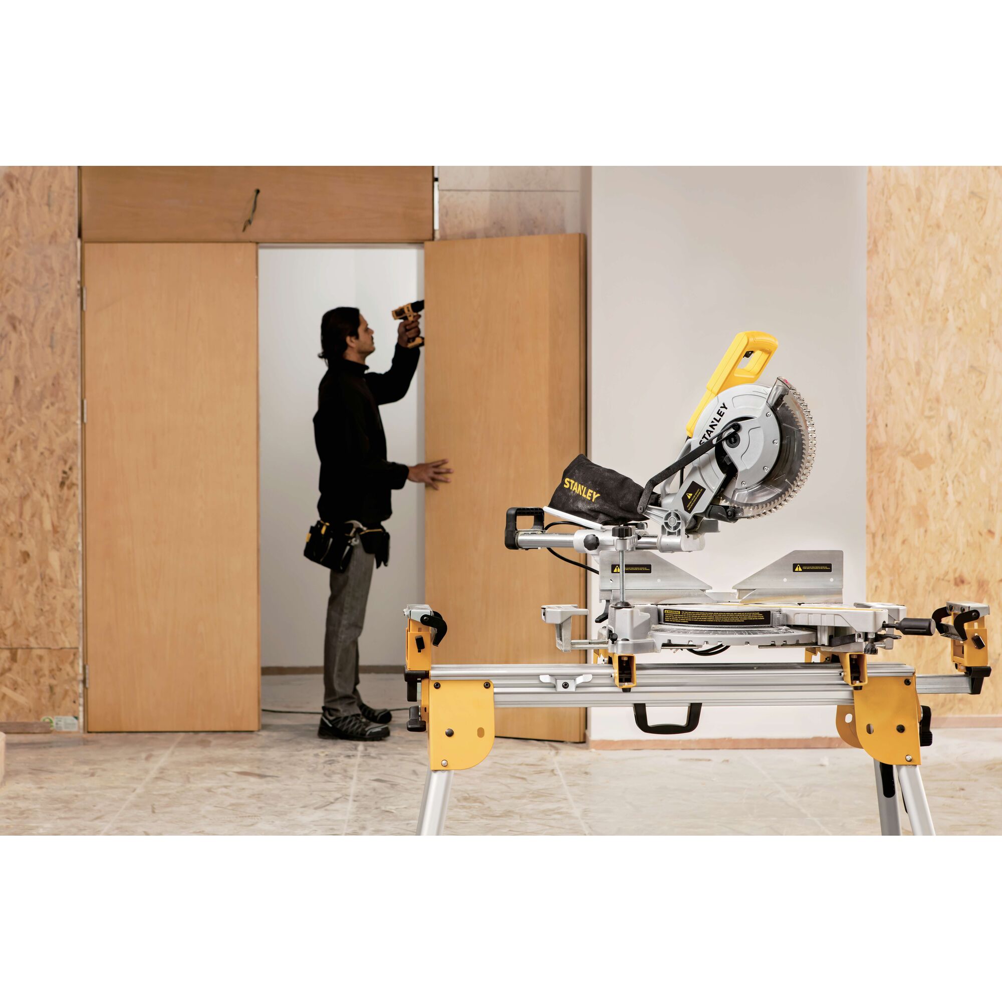 STANLEY SM18-B2C miter saw on stand in workshop, person using drill near wooden door in background.