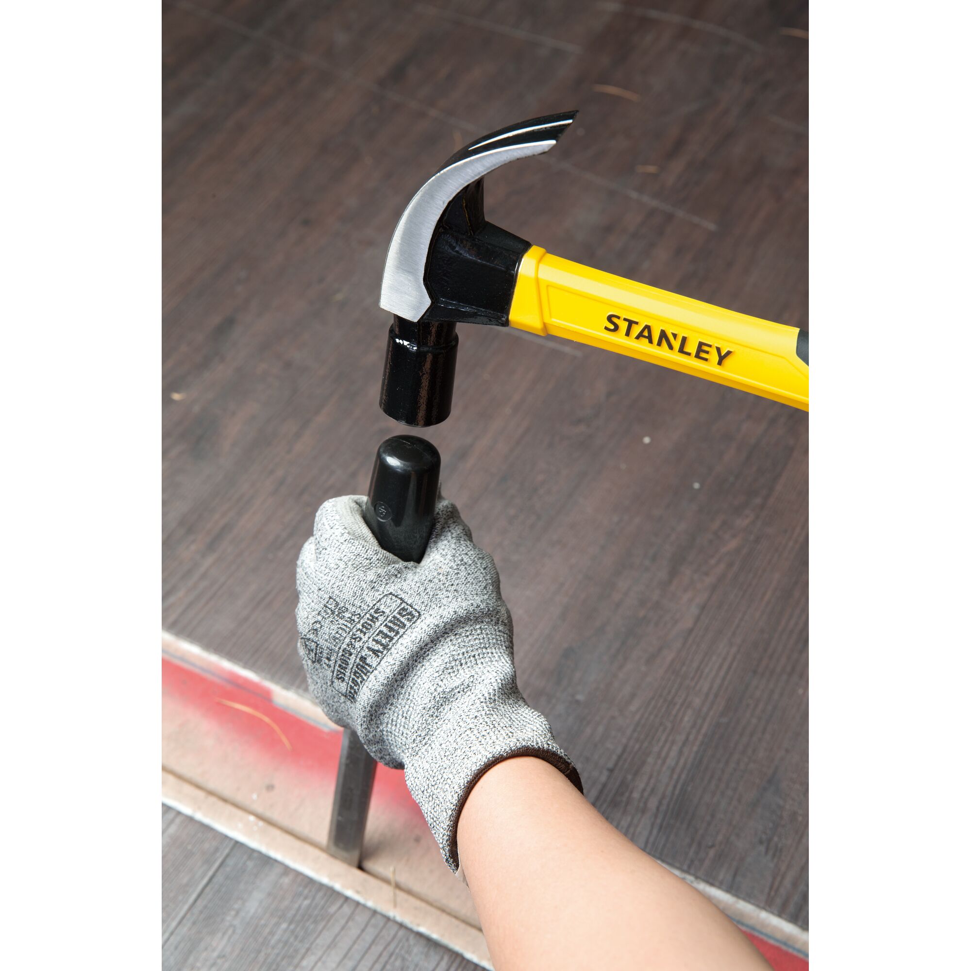 Photo of STANLEY STHT51391 claw hammer with yellow handle in use, striking a black post, person's gloved hand holding post.