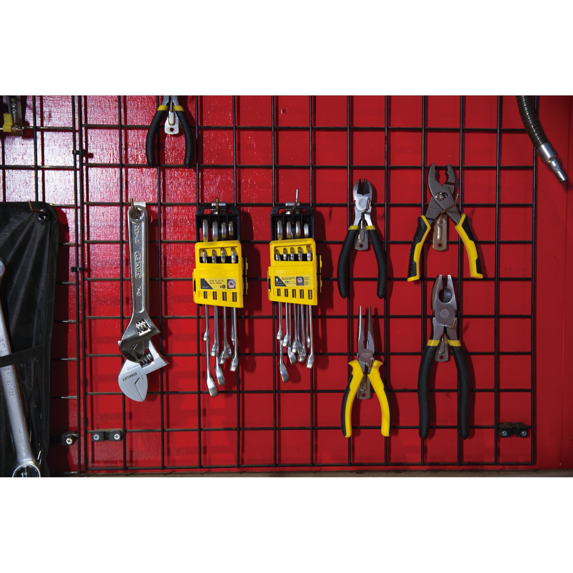 A set of hand tools including wrenches with yellow holders and various pliers with black and yellow handles are organized on a red grid wall panel.